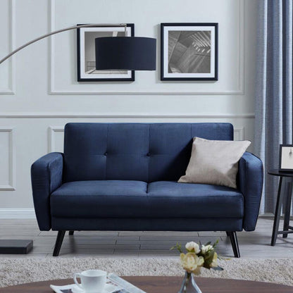 Esben 2-Seater Velvet Sofa Bed in Navy Blue with slim black-metal legs, features a light-beige lumbar pillow. Accompanied by monochrome prints, black floor lamp, side table, and low coffee table.