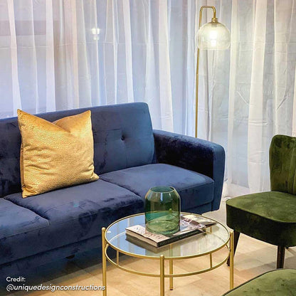 Esben 2-Seater Velvet Sofa Bed in Navy Blue, featuring a deep-blue velvet upholstery with an oversized metallic gold throw pillow, set in a stylish living room with sheer white drapes and a glass coffee table.