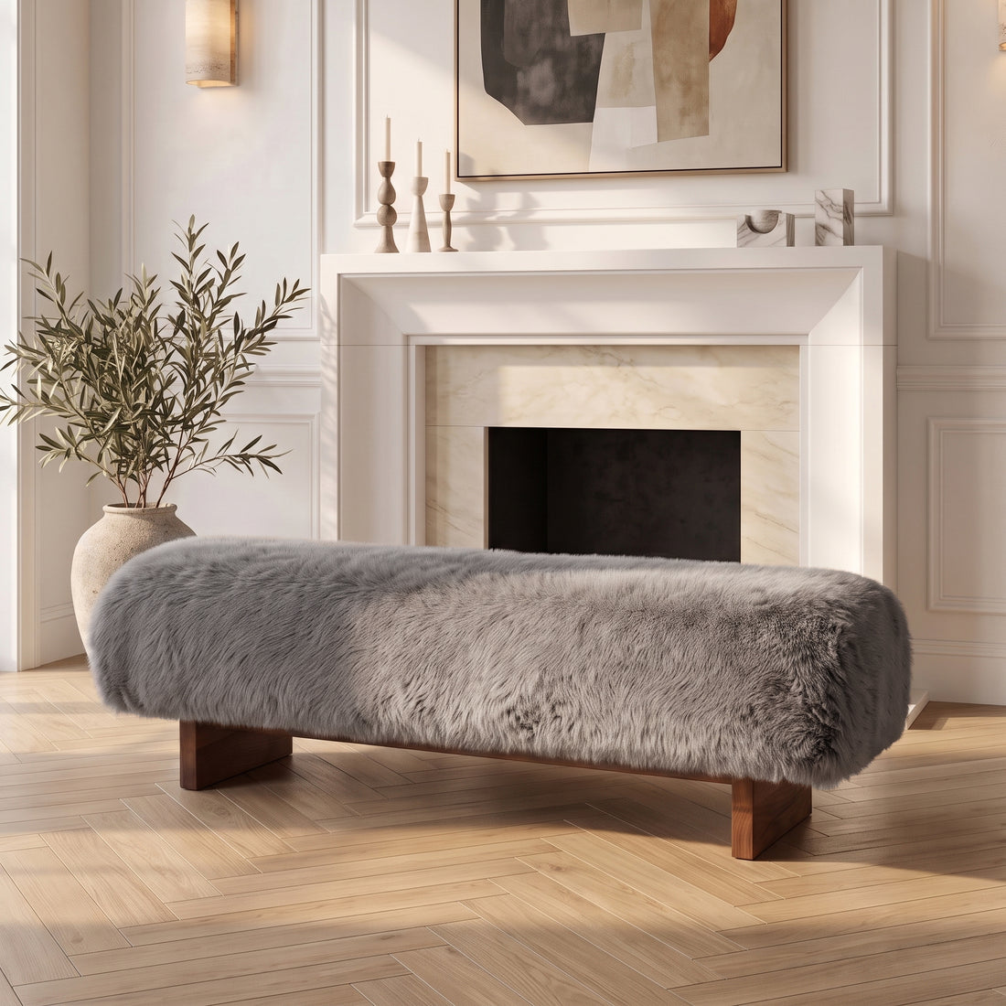 A modern, luxurious bench featuring a plush, textured gray faux-fur or sheepskin upholstered seat, supported by low, dark wooden block legs, positioned on a light herringbone wood floor in front of a classic white fireplace mantle.