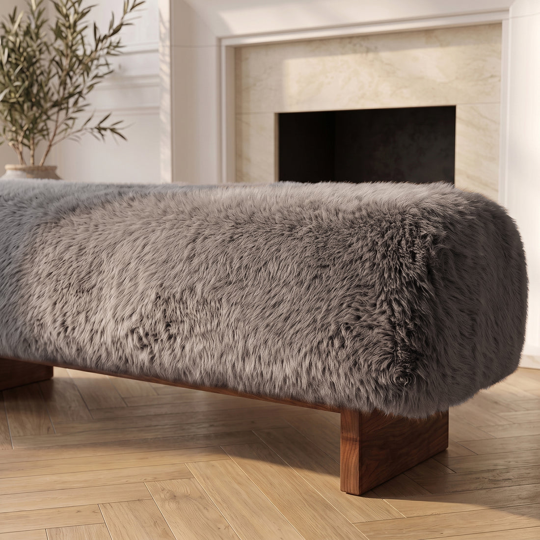 A close-up, angled view of the taupe faux-fur or sheepskin upholstered bench, highlighting its plush, long-pile texture and low dark wooden block legs, positioned on a light herringbone wood floor in a sunlit room with a classic fireplace mantle in the background.