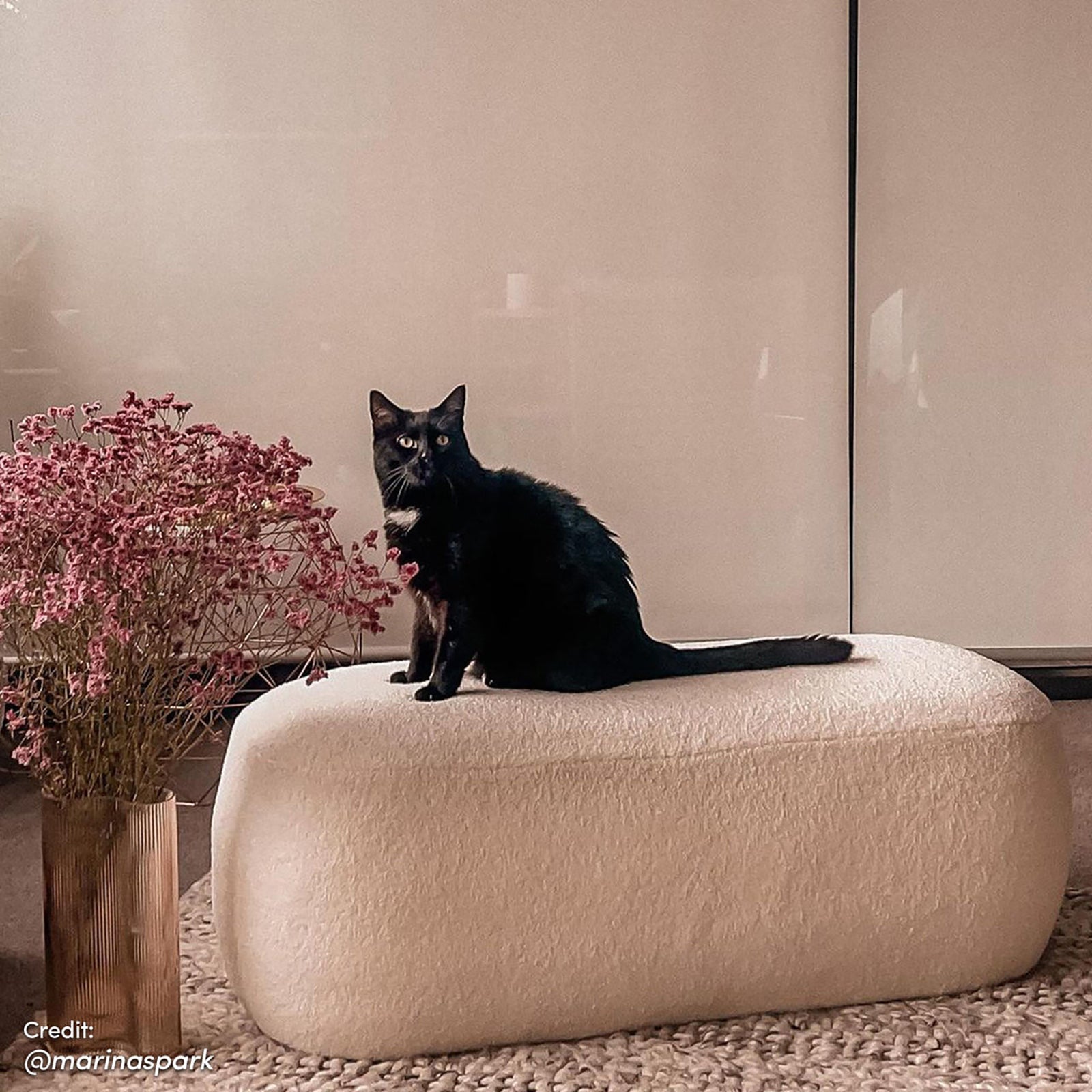 Jamie 110cm Bouclé Ottoman in Cream White with a sleek black cat on top, next to a ribbed metallic vase with dusty-pink blooms, set on a chunky-woven rug.