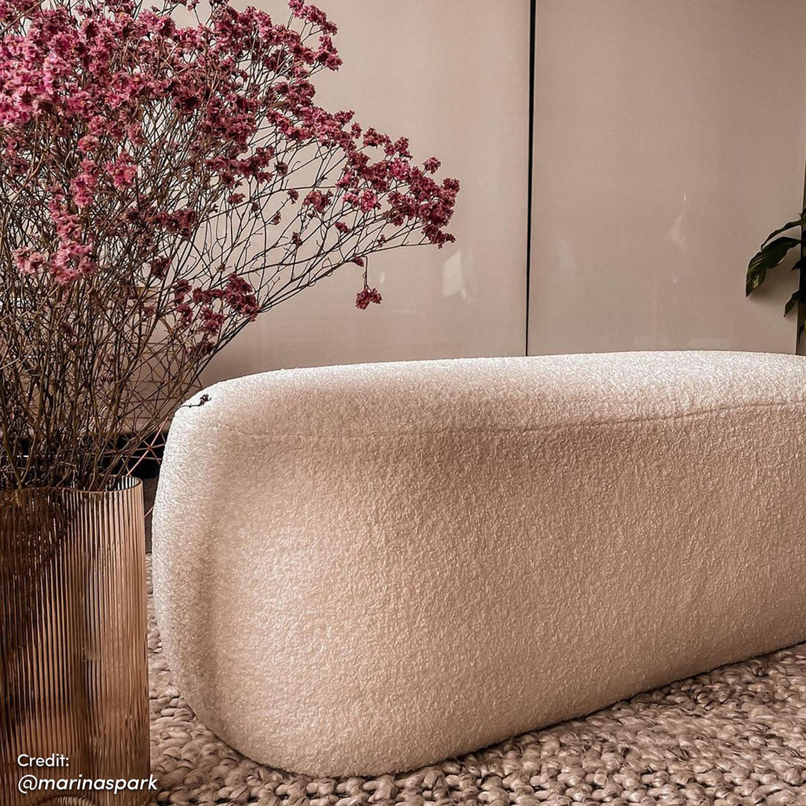 Jamie 110cm Bouclé Ottoman in Cream White on a woven rug, accompanied by a fluted metallic vase with dried pink flowers, set against smooth cabinetry and a hint of greenery.
