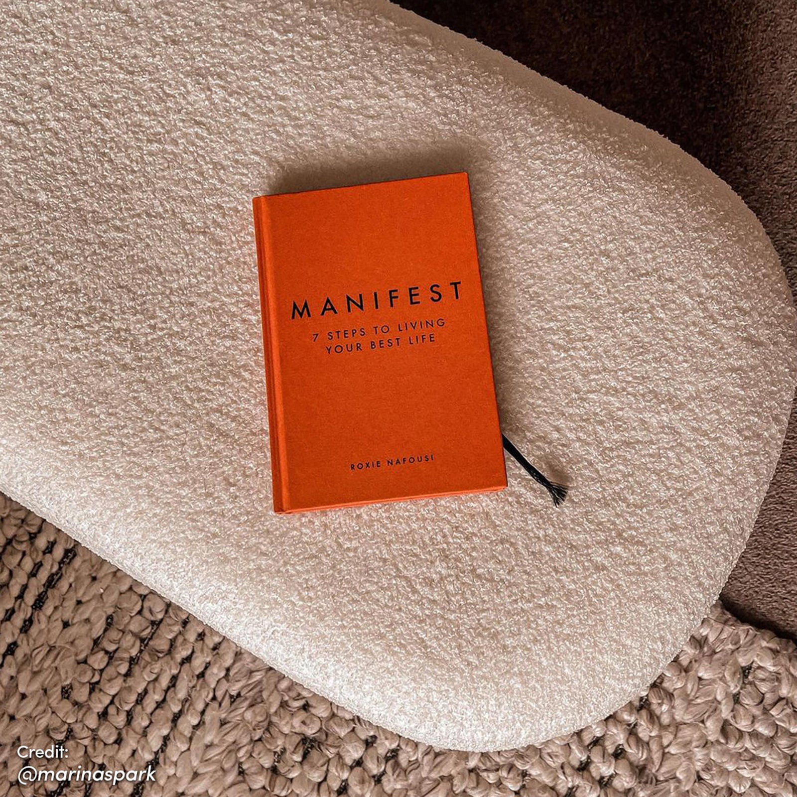 Jamie 110cm Bouclé Fabric Ottoman in Cream White, topped with a bright orange hardcover journal titled MANIFEST by Roxie Nafousi, rests on a chunky woven rug with a black ribbon bookmark visible.