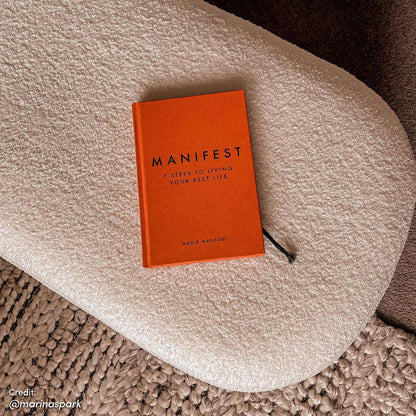 Jamie 110cm Bouclé Fabric Ottoman in Cream White, topped with a bright orange hardcover journal titled MANIFEST by Roxie Nafousi, rests on a chunky woven rug with a black ribbon bookmark visible.