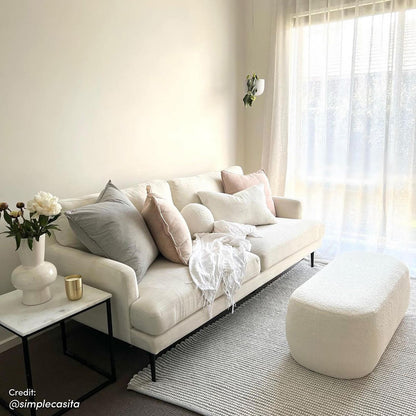 Jamie 110cm Bouclé Ottoman in Cream White, featuring a rounded shape and rich texture. Positioned on a light-gray rug, it complements a cream-colored sofa and adds elegance to the living room setting.