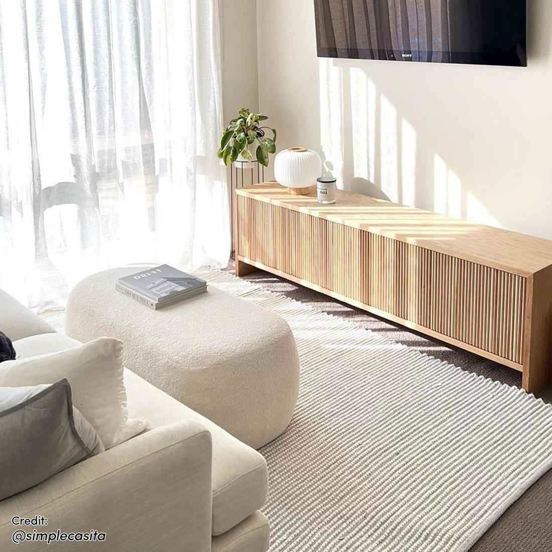 Jamie 110cm Boucle Ottoman in Cream White, a kidney-shaped ottoman with soft texture, topped with a book, nestled in a bright living room corner, offering a cozy, elegant resting space.