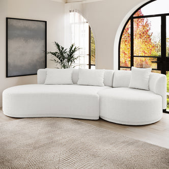 Lyana 4 Seater Boucle Sofa with Left Chaise in Cream White, featuring a curved design, three pillows, and positioned on a geometric rug with sunlight through arched windows.