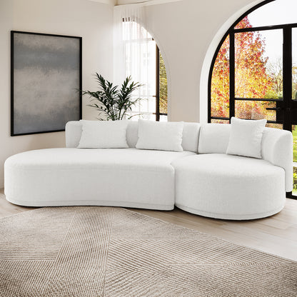 Lyana 4 Seater Boucle Sofa with Left Chaise in Cream White, featuring a curved design, three pillows, and positioned on a geometric rug with sunlight through arched windows.