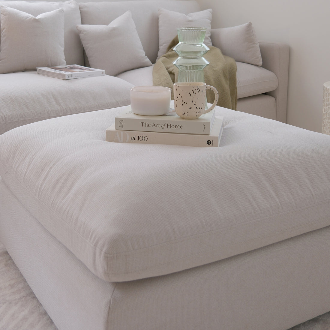 Millie Fabric Ottoman Modular Sofa in warm light grey shown with a white pillow, spotted mug, and book, highlighting its plush, deep seating, and versatile design for contemporary living spaces.