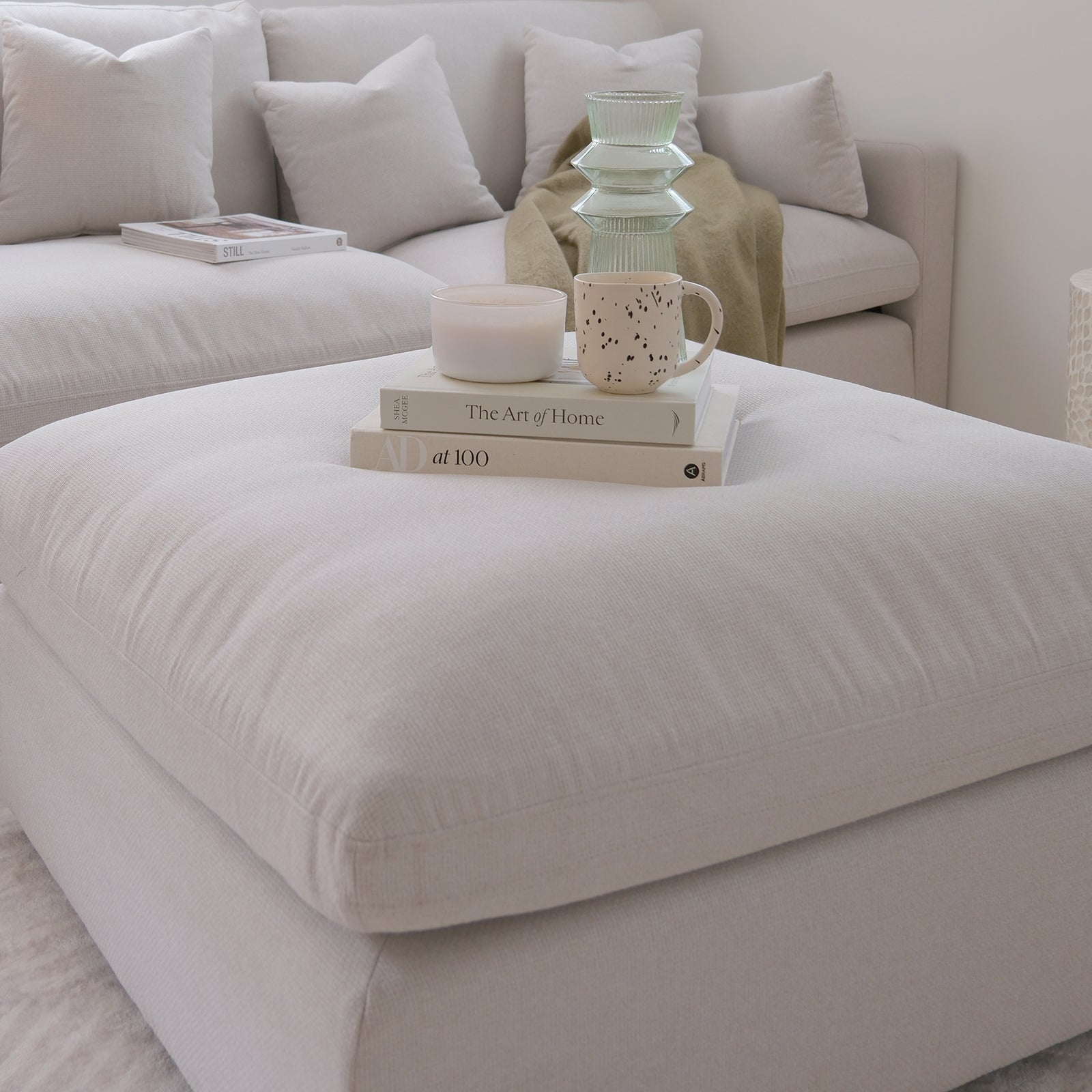 Millie Fabric Ottoman Modular Sofa in warm light grey shown with a white pillow, spotted mug, and book, highlighting its plush, deep seating, and versatile design for contemporary living spaces.