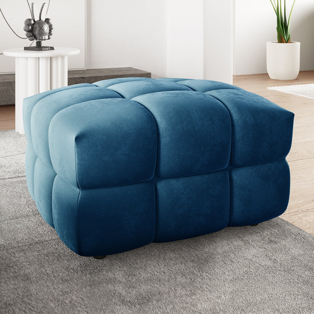 Norah Velvet Upholstered Tufted Ottoman in Blue, showcasing a plush, grid-tufted design on a light gray rug, with a modern living-room backdrop featuring a white side table and potted plant.