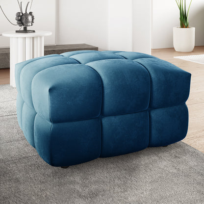 Norah Velvet Upholstered Tufted Ottoman in Blue, showcasing a plush, grid-tufted design on a light gray rug, with a modern living-room backdrop featuring a white side table and potted plant.