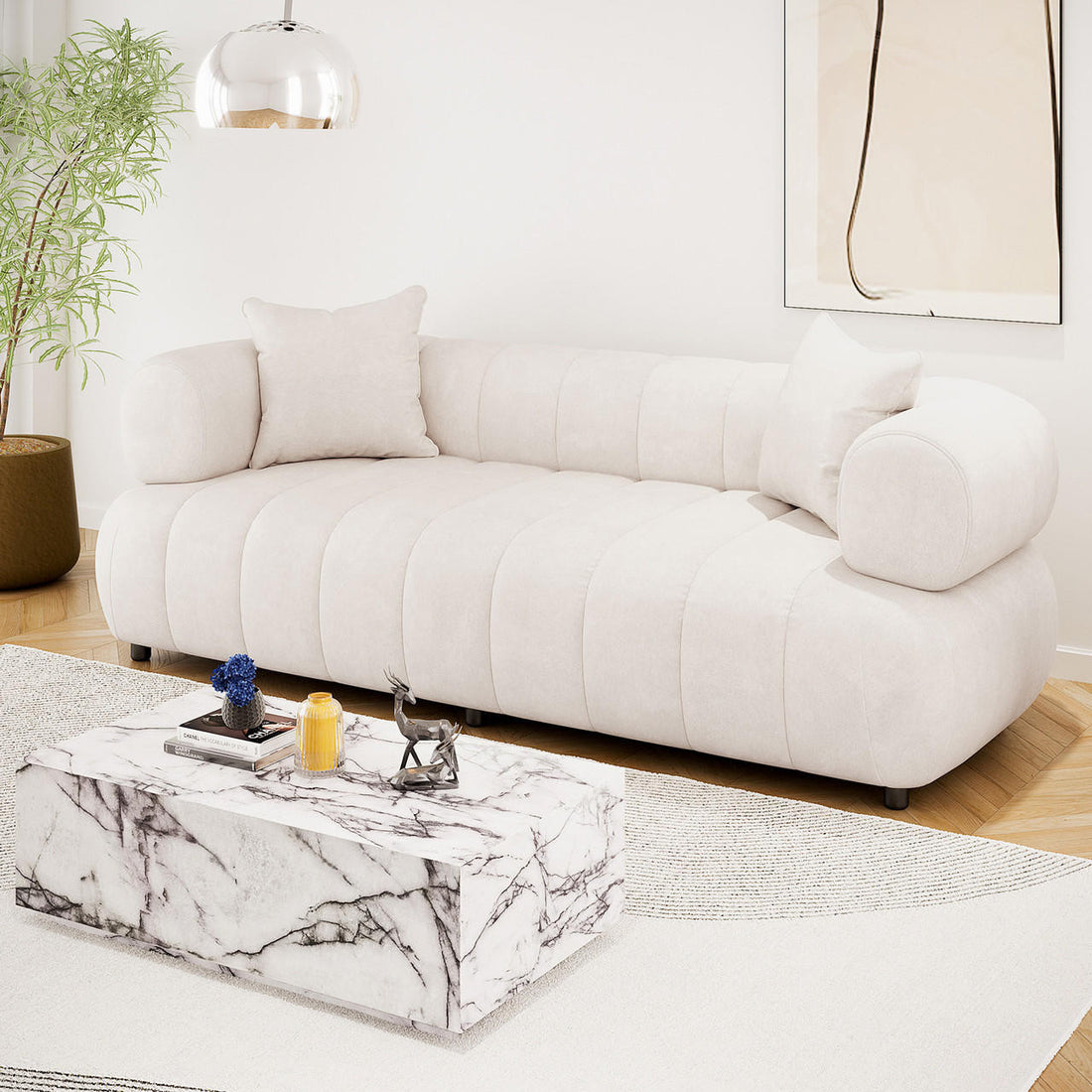 Samira 3 Seater Velvet Sofa in Cream with rounded arms and two matching pillows, paired with a marble-patterned coffee table and decorative items, set in a stylish and serene living room.