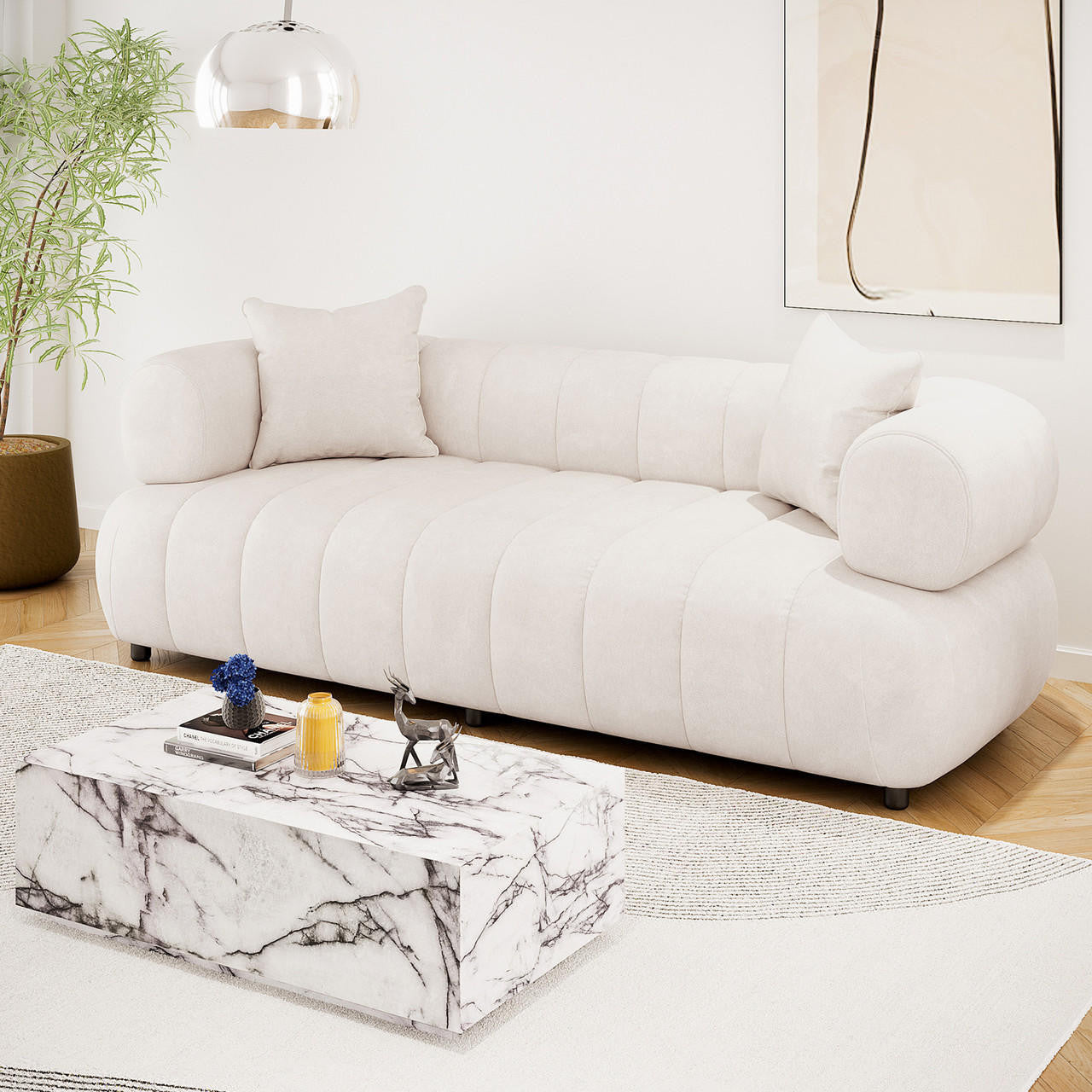 Samira 3 Seater Velvet Sofa in Cream with rounded arms and two matching pillows, paired with a marble-patterned coffee table and decorative items, set in a stylish and serene living room.