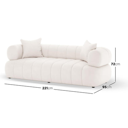 Samira 3 Seater Velvet Sofa in Cream features a low-profile design with rounded arms, vertical channel-tufted panels, and two throw pillows, offering plush comfort and sophisticated style.