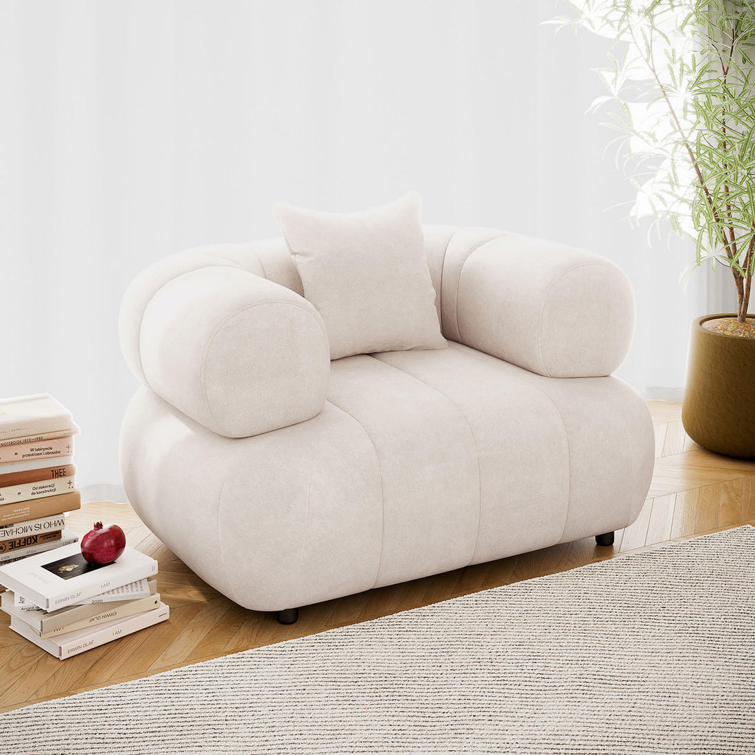 Samira Single Seater Velvet Sofa in Cream features an oversized design with chunky, tubular arms and a matching throw pillow, set on a herringbone-patterned wood floor with books nearby.