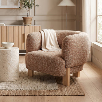 Sornel Boucle Upholstered Accent Chair in Coral with rounded design and boucle fabric, wooden legs, beside a white side table and light-wood cabinet with plant, enhancing cozy living space ambiance.
