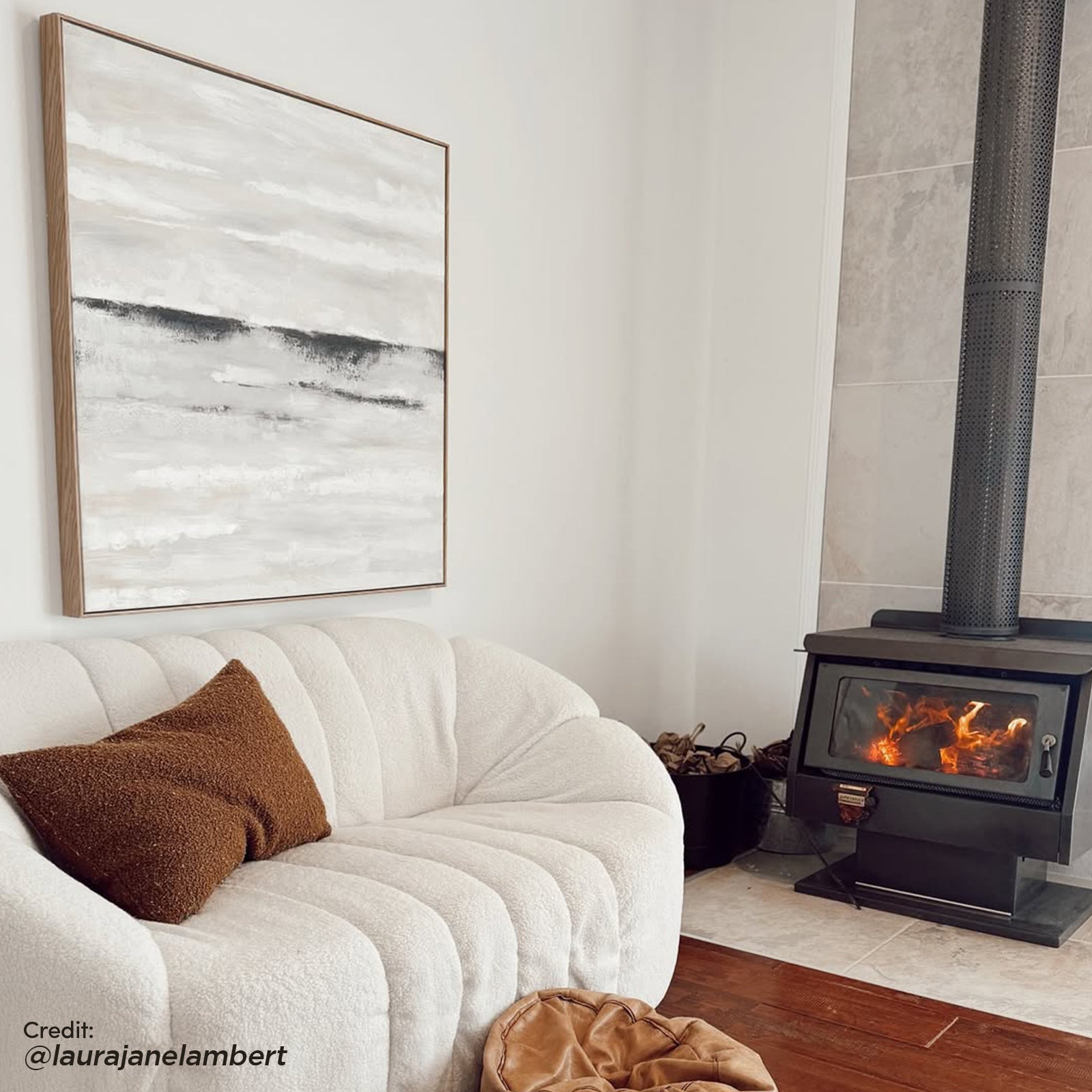 Trina 3 Seater Boucle Sofa with Pillows in Cream White, featuring plush channel-tufted upholstery, a rust-brown throw pillow, and a cozy setting with a wood-burning stove and hardwood flooring.