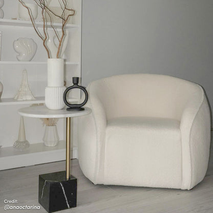 Yara Bouclé Accent Chair in Cream White, featuring a low, rounded design with textured upholstery, next to a marble-topped side table with decorative ceramic vase and branches, set in a minimalist living space.