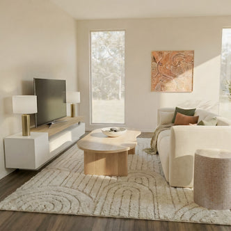 Organic modern living room featuring a cream-colored sofa with earth-tone pillows, a light oak kidney-shaped coffee table, and a textured cream area rug. A large wall-mounted flat-screen TV sits on a white and oak minimalist entertainment unit flanked by modern globe lamps.