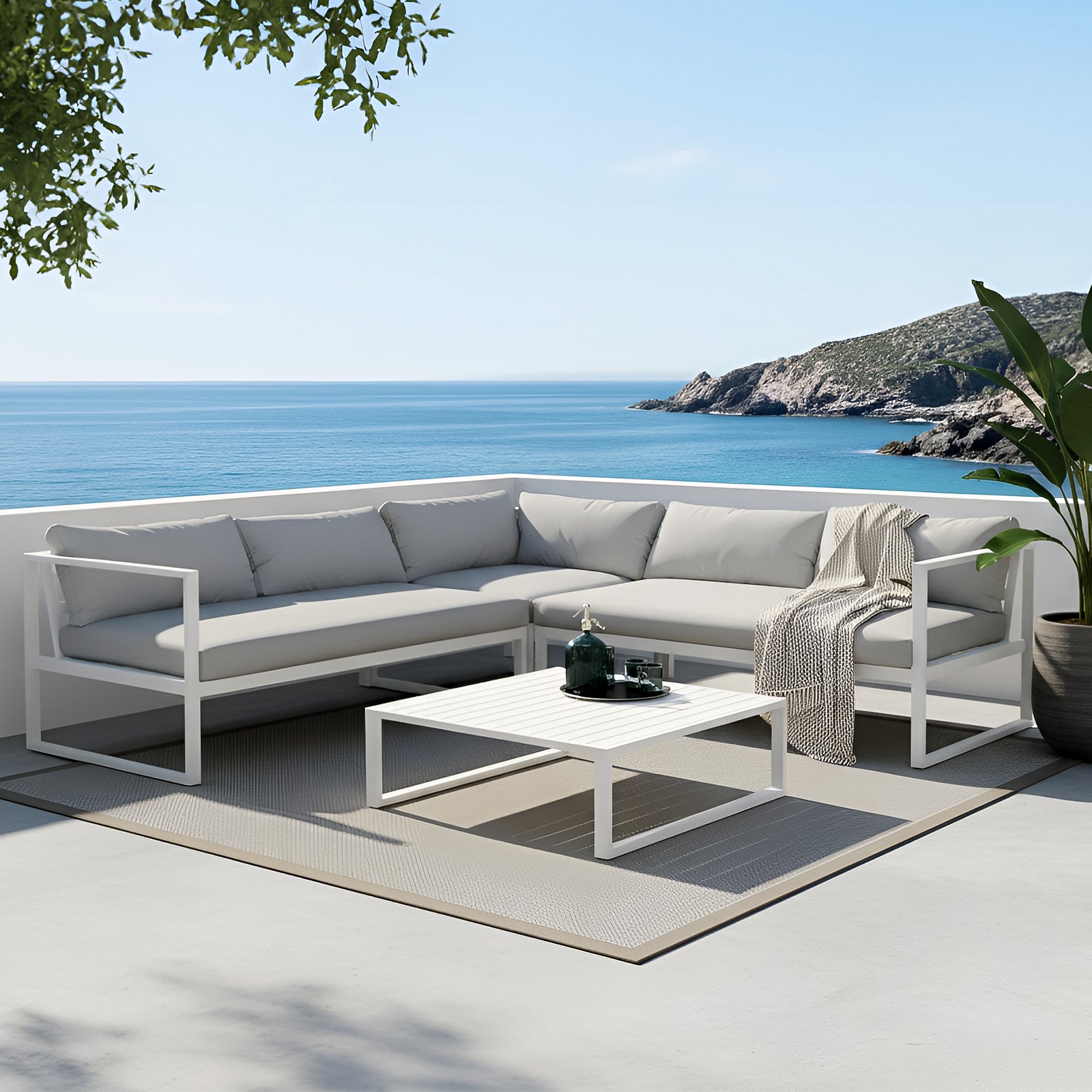 Minimalist outdoor patio set featuring a large white aluminum L-shaped sectional sofa with grey weather-resistant cushions. A matching white slatted coffee table sits on a grey outdoor rug against a Mediterranean coastal backdrop with blue ocean views and a potted bird of paradise plant.