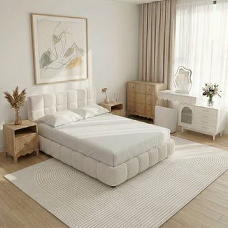 A modern bedroom featuring a tufted cream bouclé bed, light wood bedside tables, a white vanity with a wavy mirror, and a linear-patterned rug.