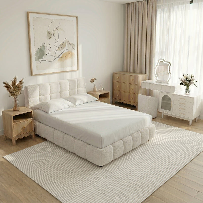 Cozy, light-filled nursery with Scandinavian style decor. A sage-green crib with scalloped edges sits against a white wall beneath three minimalist framed art prints. To the left is a matching green dresser with a small lamp and children’s books on top. To the right, a boucle lounge chair with a rust throw blanket and a matching ottoman with wooden legs face a large window. Shallow wall shelves display wooden toys and picture books. A round cream rug with colorful polka dots lies on pale wood floors. The room feels calm, tidy, and child-friendly.