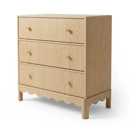 A light oak dresser featuring three spacious drawers, each accented with gold flower-shaped knobs, and finished with a scalloped base detail.