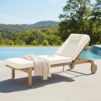 Zyra Hardwood Reclining Sun Lounger with Cushion in Cream, featuring a wooden frame, adjustable backrest, cream cushion, knit throw, and sunglasses by an infinity pool with scenic hills.
