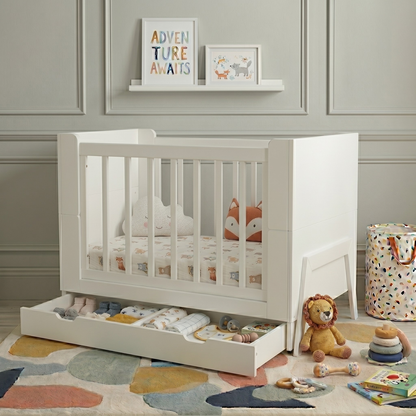 White crib in nursery corner with plush toys, storage drawer and geometric rug