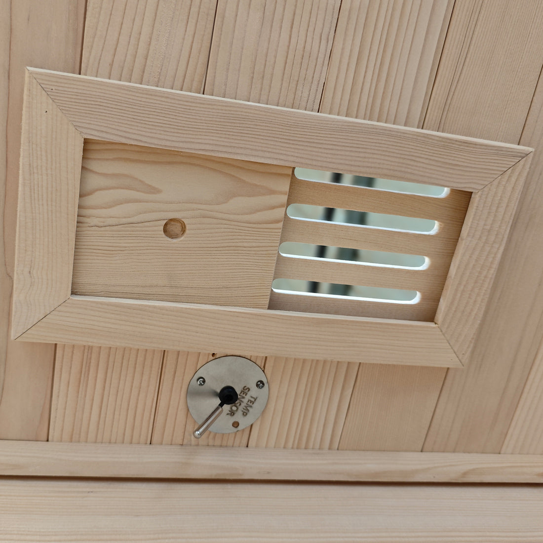 Wooden vent cover with a round metal control dial on a wood-paneled surface.