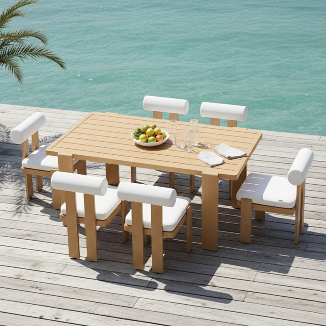 Positano 6 Seater Aluminium Outdoor Dining Set in White on a wooden deck with fruit bowl, glass carafes, and white-cushioned chairs, overlooking turquoise water beneath palm fronds.
