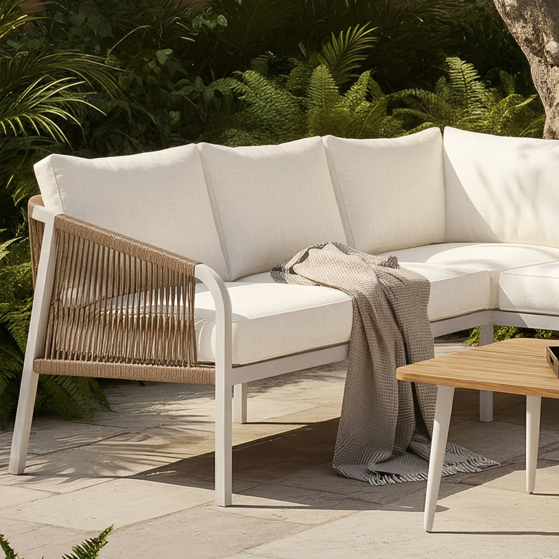 Outdoor lounge with Vernon Sofa Cushion Covers in Cream White on a curved sectional. Features woven rope frame and textured gray throw. Lush foliage backdrop enhances the tranquil setting.