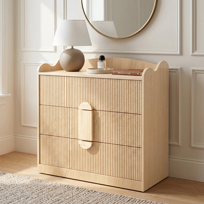 Three-drawer wooden dresser in a bedroom setting with lamp, mirror, and decor