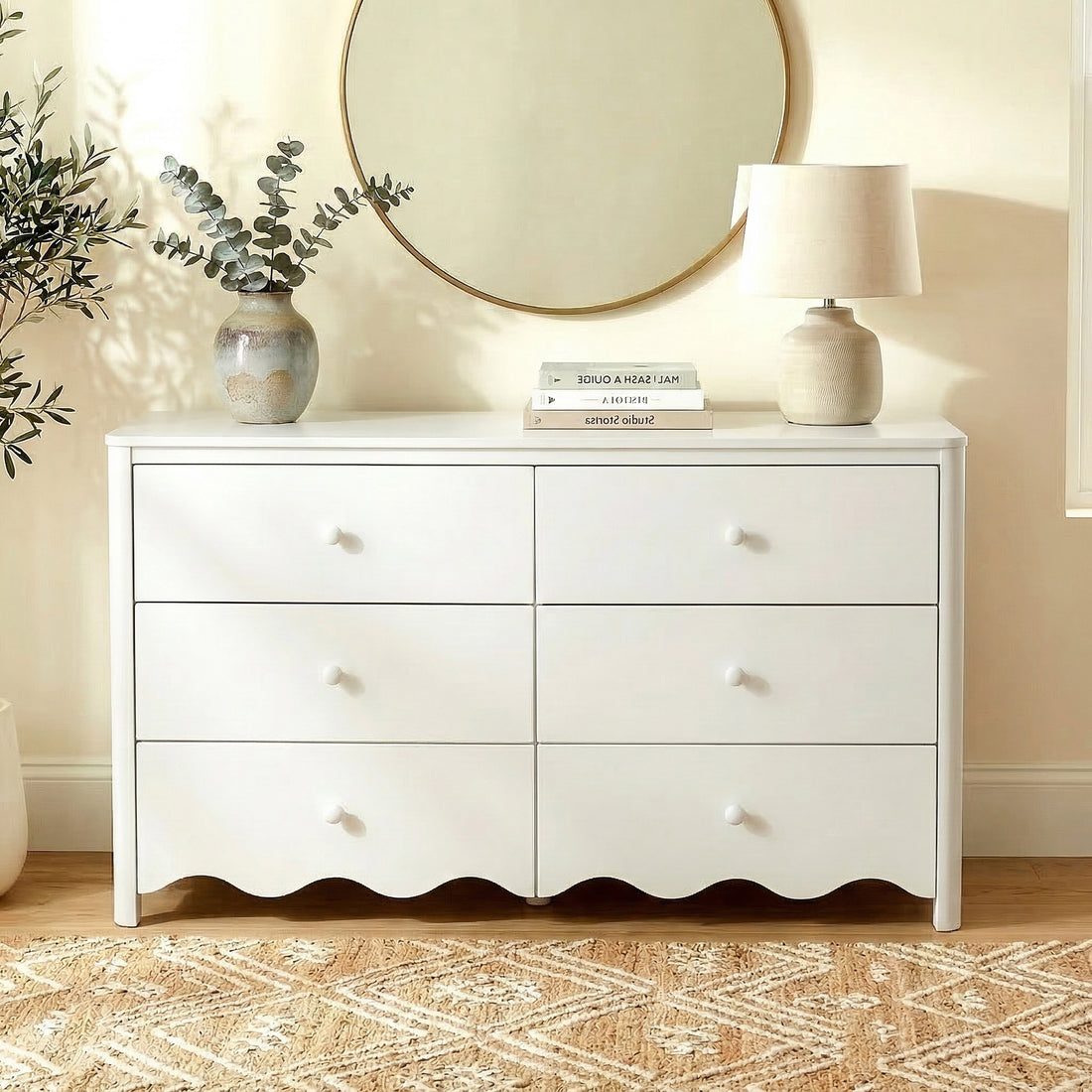 White dresser in a softly lit room with vase, books, lamp, and mirror
