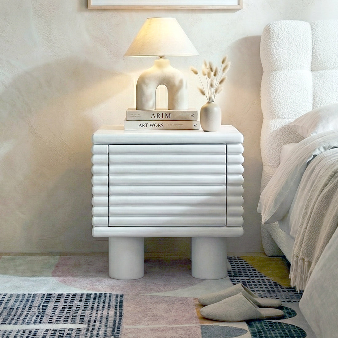 White ribbed nightstand styled in a cozy bedroom with lamp, books, vase and slippers
