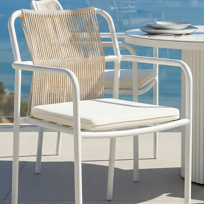 Evone Outdoor Cushion Covers in Cream White adorn bar-height chairs with woven rope backs, enhancing the coastal vibe of a seaside setting with a round white table and ocean backdrop.