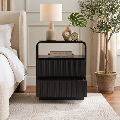 Black nightstand styled beside a bed in a neutral bedroom with lamp, books, and decor