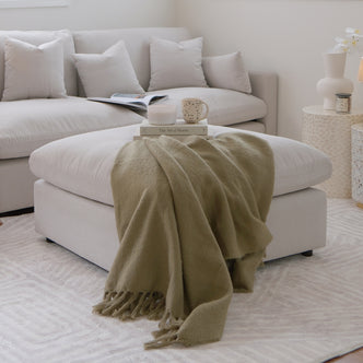 Light beige ottoman styled in a modern living room with sectional sofa, books, and decor