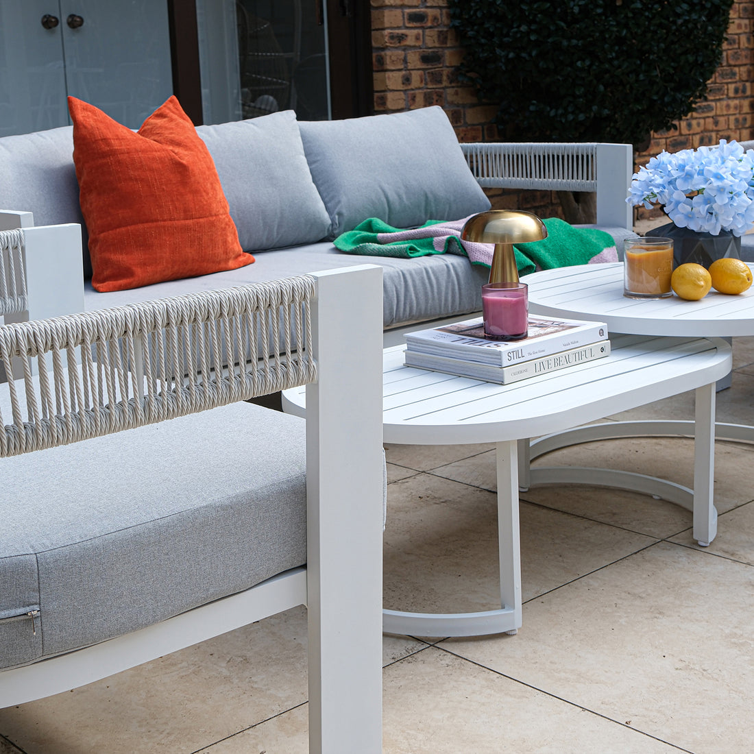 Outdoor patio scene featuring a light grey sectional sofa with orange pillow, green throw, and styled coffee table