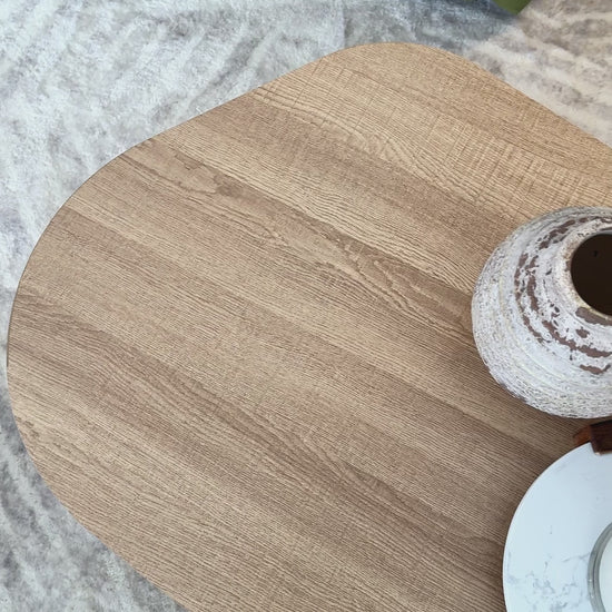 
A close-up of a modern, light oak coffee table with a freeform, organic shape. The camera slowly pans over the table's surface, showing its natural wood grain. The table is styled with two rustic vases and a lit candle on a marble tray. The shot also shows a plush, gray rug and the corner of a green velvet sofa, highlighting the cozy, inviting aesthetic of the living space.