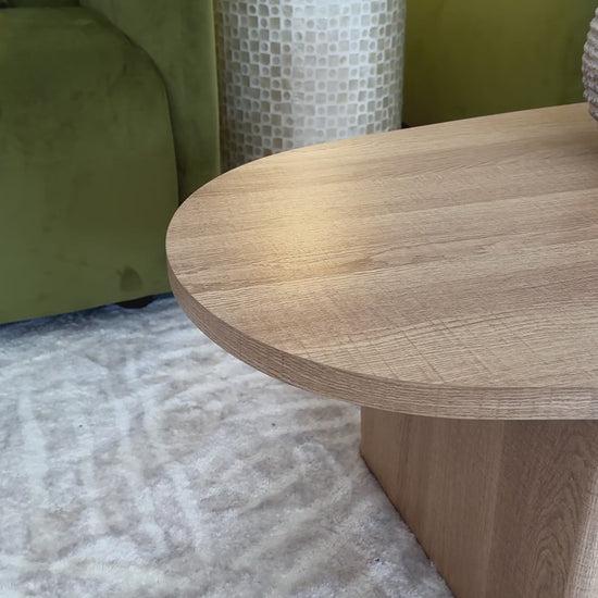 
A slow, cinematic close-up of a modern living room coffee table, showcasing its freeform, organic shape and light oak wood finish. The tabletop's natural grain is visible as the camera pans slowly across it. The table is styled with a rustic-looking vase, a larger textured vase, and a lit candle on a marble tray. The shot then pans up to show a vibrant green velvet sofa with pillows and a side table with a decorative lamp, creating a cozy and inviting atmosphere.